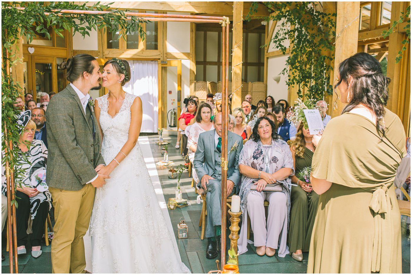 Bride and Groom stand under copper arch way as bridesmaid gives a reading