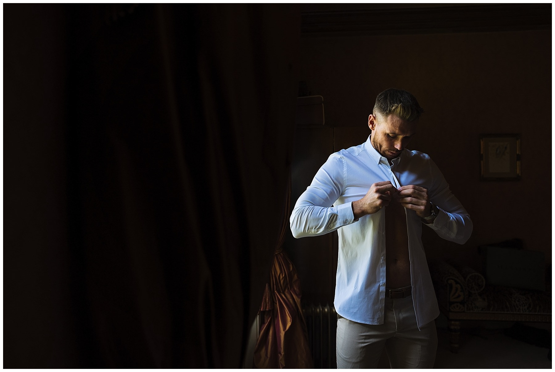 groom buttons his shirt in the window