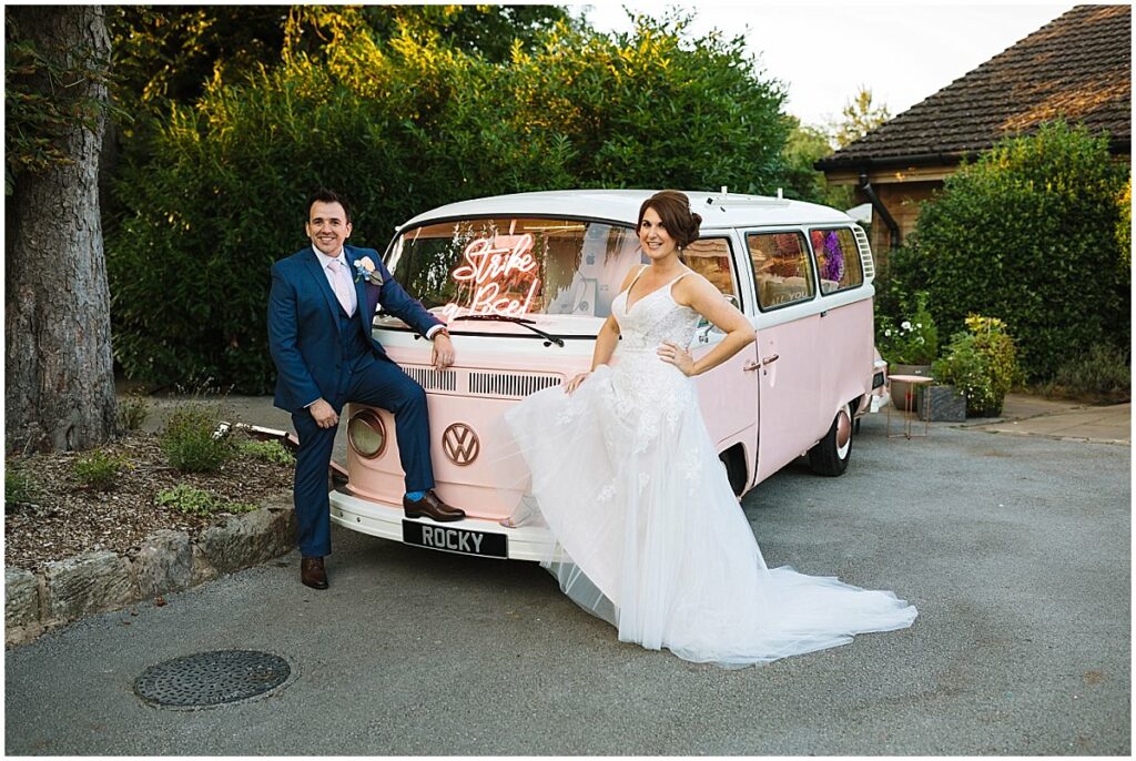 Bride and Groom stand in front of Bright Pink Camper Van With ROCKY number plate
