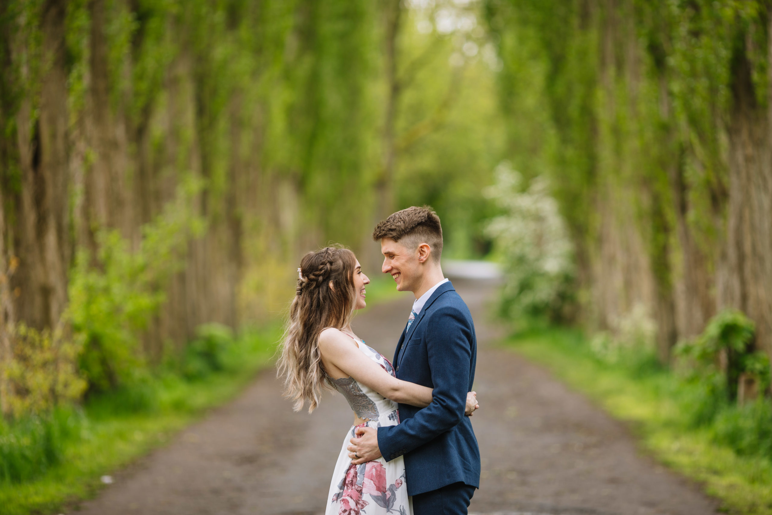 A happy couple in semi-formal attire embracing and smiling at each other on a tree-lined path.