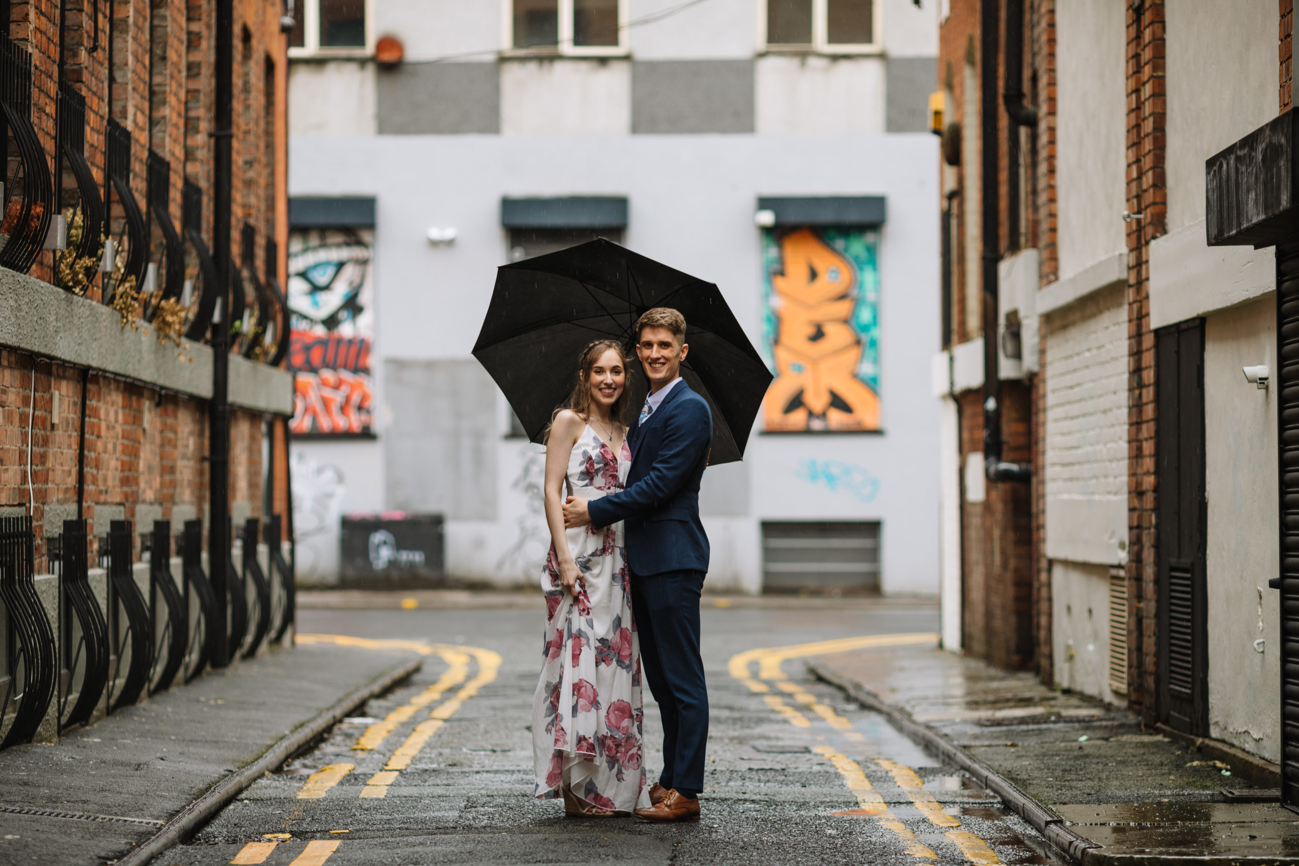 A smiling couple standing under an umbrella on a wet street with urban graffiti in the background.