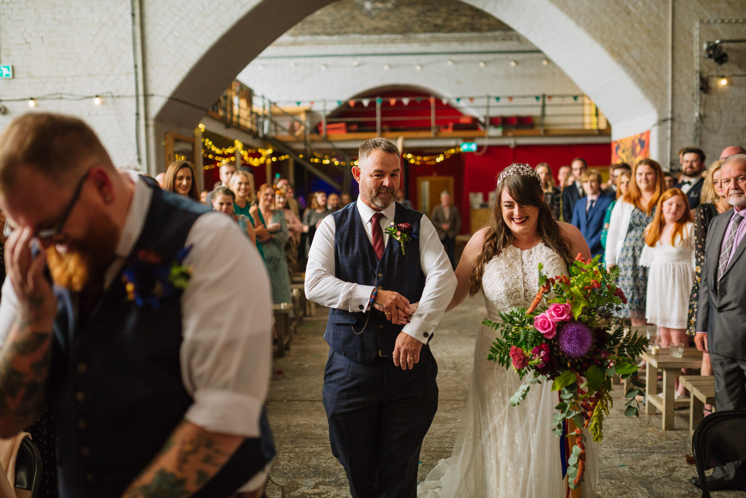 A bride with a bright bouquet and a groom, both smiling as they walk down the aisle, with guests watching from the sides in a festively decorated venue.