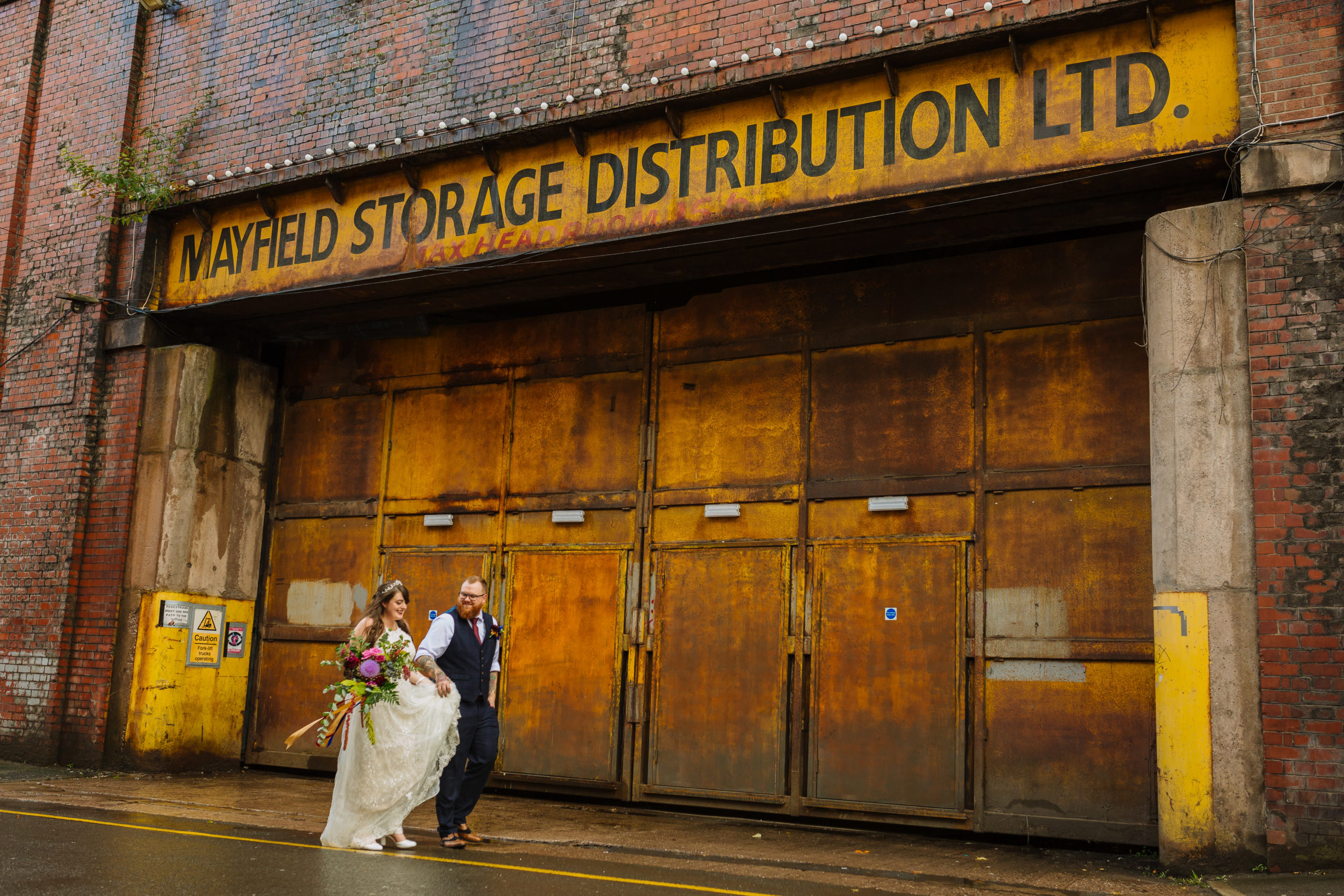A bride in a white dress and a groom in a blue suit and tie walk hand-in-hand in front of a rugged industrial backdrop with weathered orange doors and a faded sign reading "Mayfield Storage Distribution LTD."