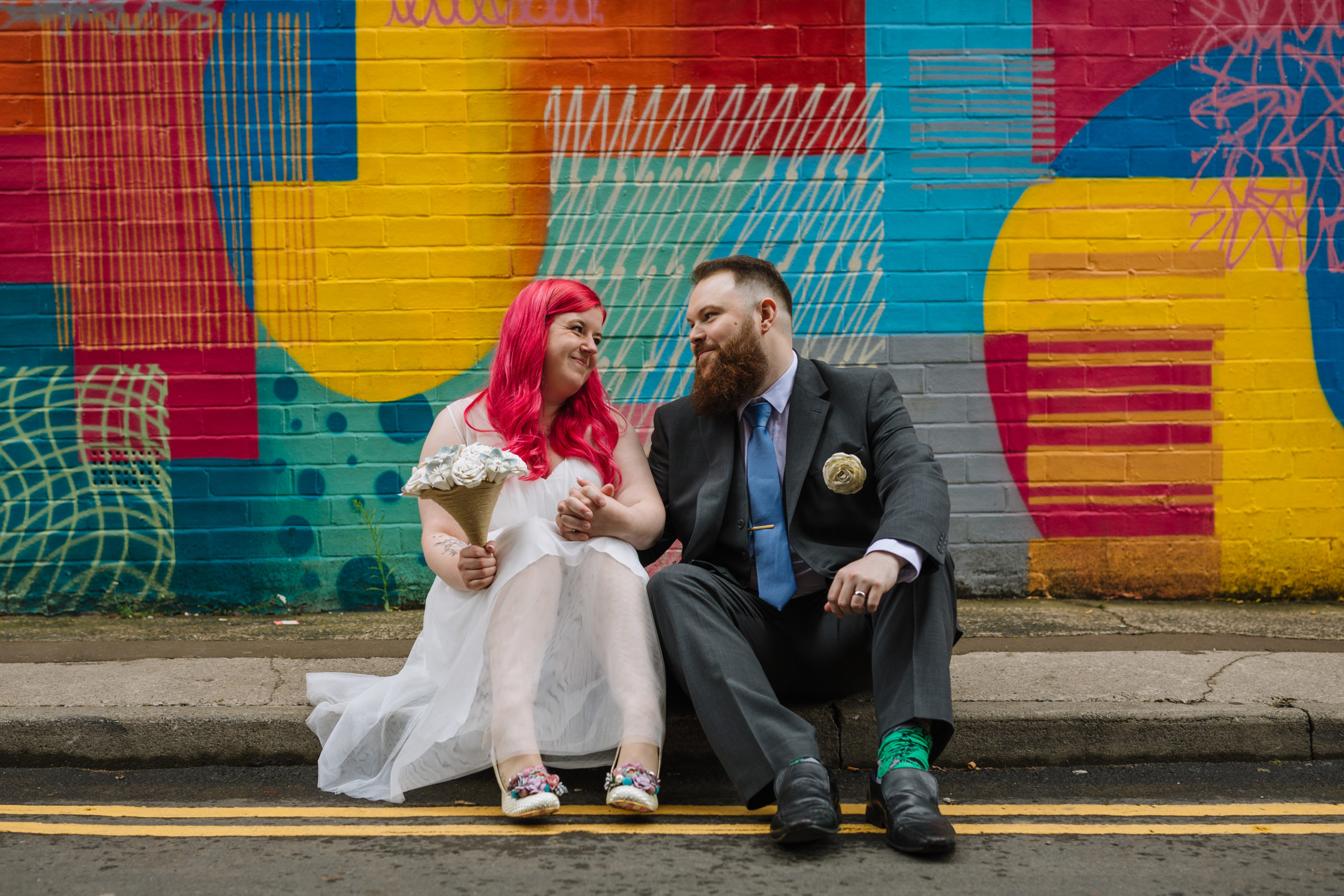 A bride with pink hair and a groom with a beard sitting on a curb, smiling at each other against a vibrant multicolored graffiti wall. The bride holds a bouquet of white flowers, and both are wearing colorful shoes.