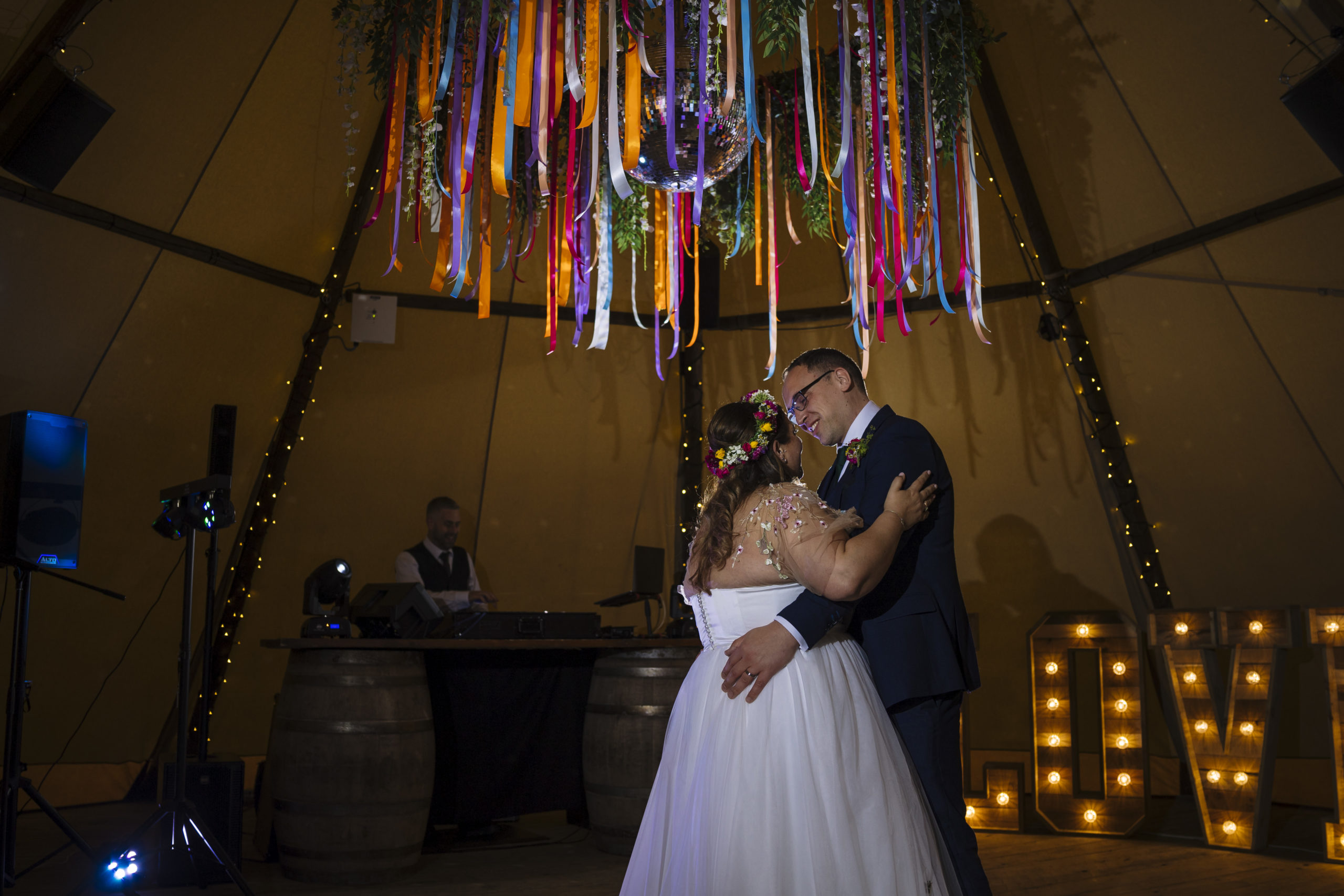 A bride and groom sharing a dance under a chandelier decorated with colorful ribbons, with a DJ booth in the background and light-up letters on the floor.