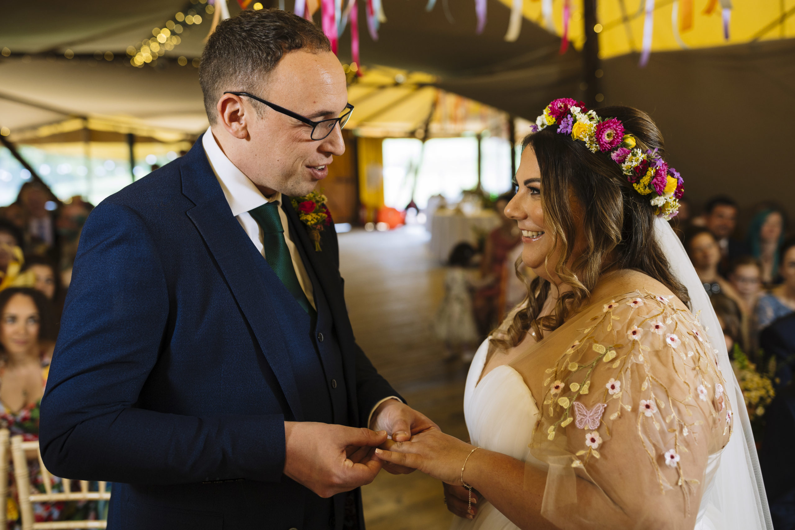 A groom in a blue suit and green tie is placing a wedding band on the bride's finger. The bride, wearing a white dress with floral embroidery and a colorful flower crown, smiles at him. They stand indoors with wedding guests in the background.