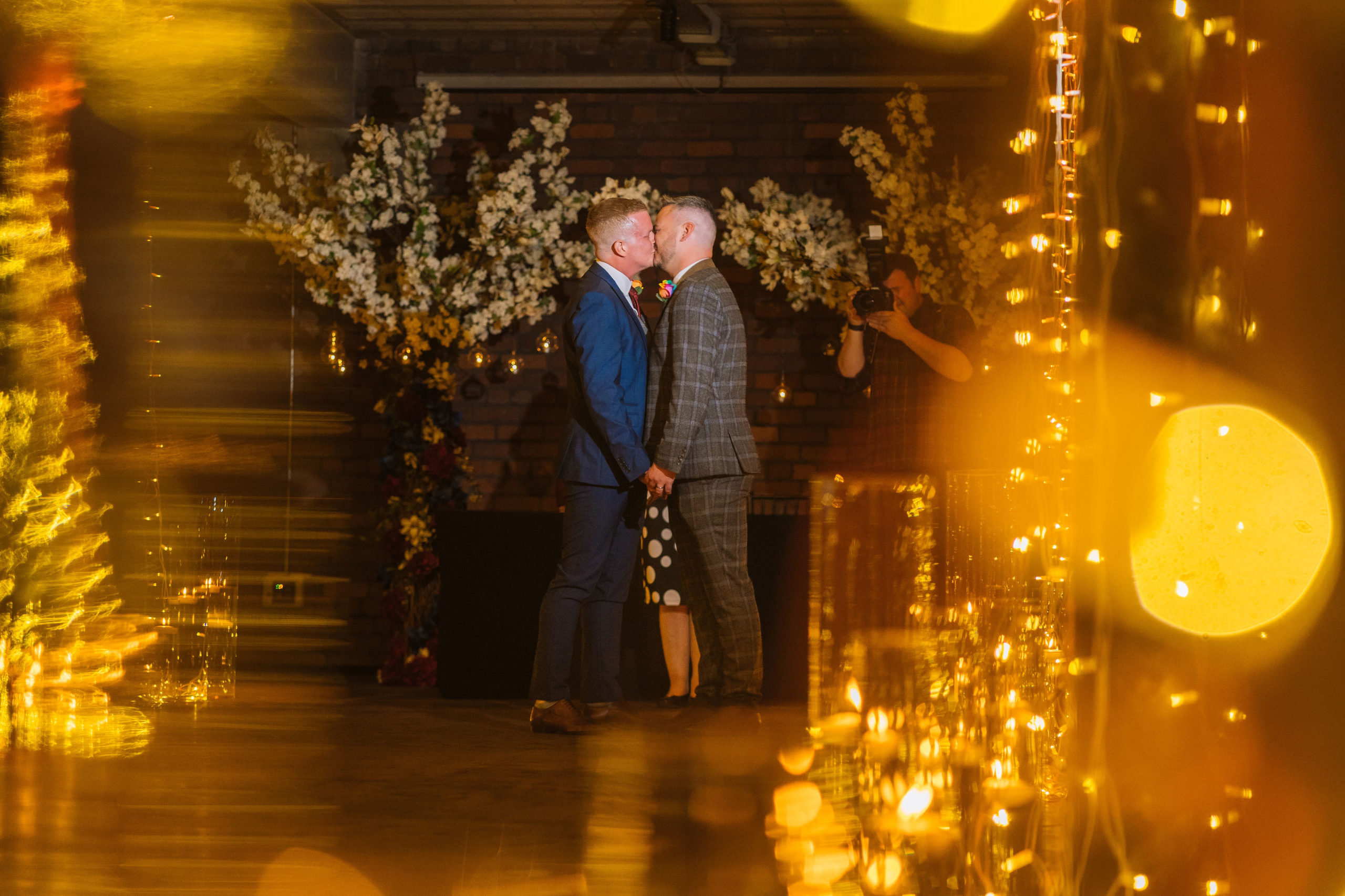 Two men kissing in a warmly lit room adorned with floral arrangements and twinkling lights, with a photographer capturing the moment in the background.