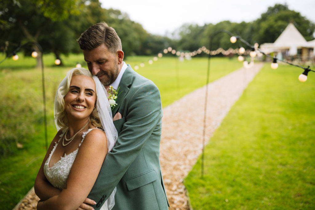 A joyful bride and groom embrace in a picturesque outdoor setting adorned with string lights and lush greenery at the tipis at riley green