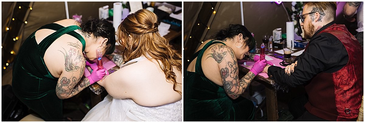 A tattoo artist in a green velvet dress tattoos a wedding guest while another guest in a white dress watches on; festive tent setting with decorations in the background.