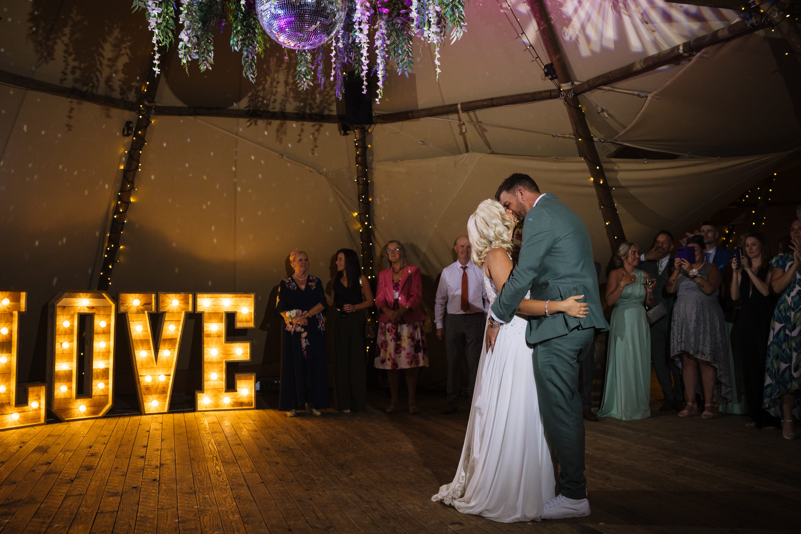 A couple shares a romantic first dance under a disco ball, surrounded by friends and family, with a large illuminated "LOVE" sign in the background.