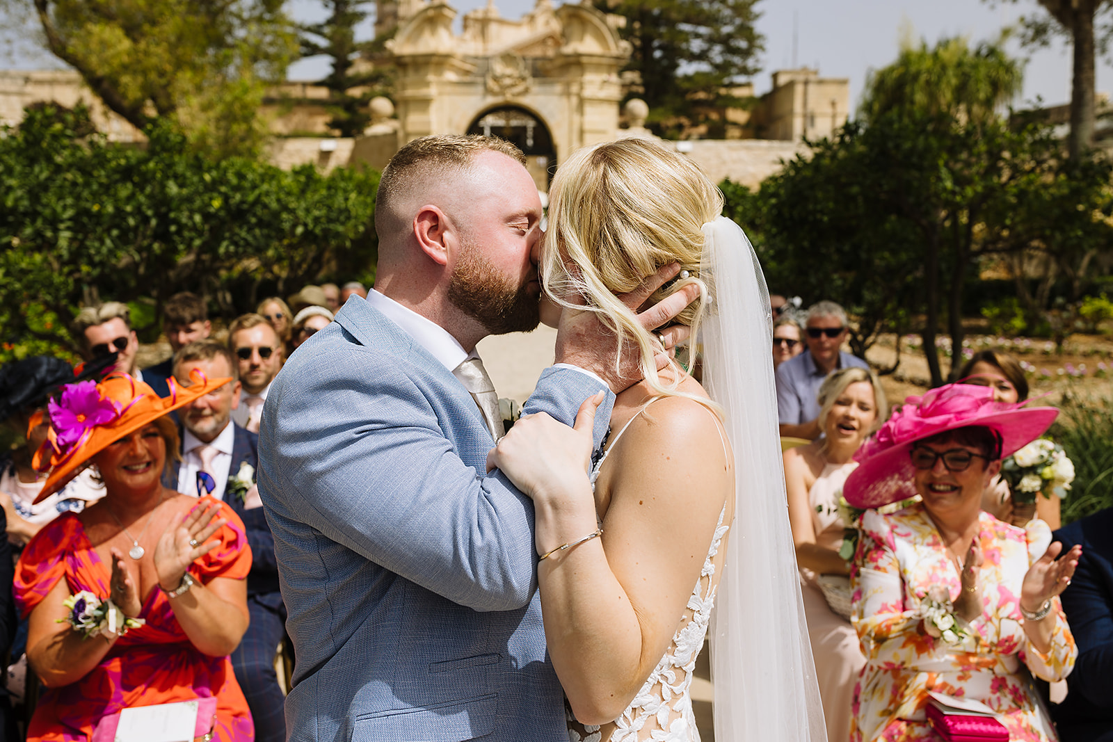 A bride and groom share a kiss during their wedding ceremony, with an applauding audience in the background and a picturesque venue setting at Palazzo Parisio in Malta