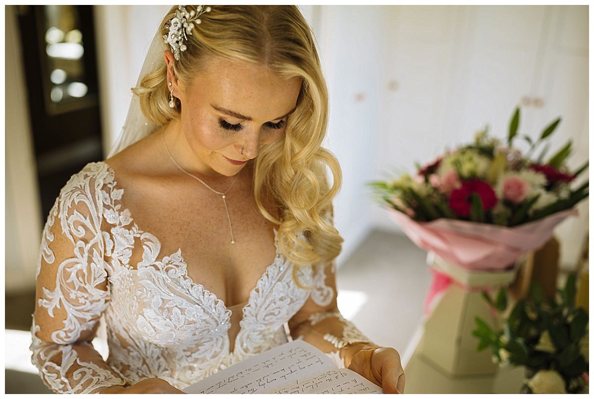 A close-up of a bride in an intricately designed lace wedding dress, focused on reading a letter, with a bouquet of flowers in the background.