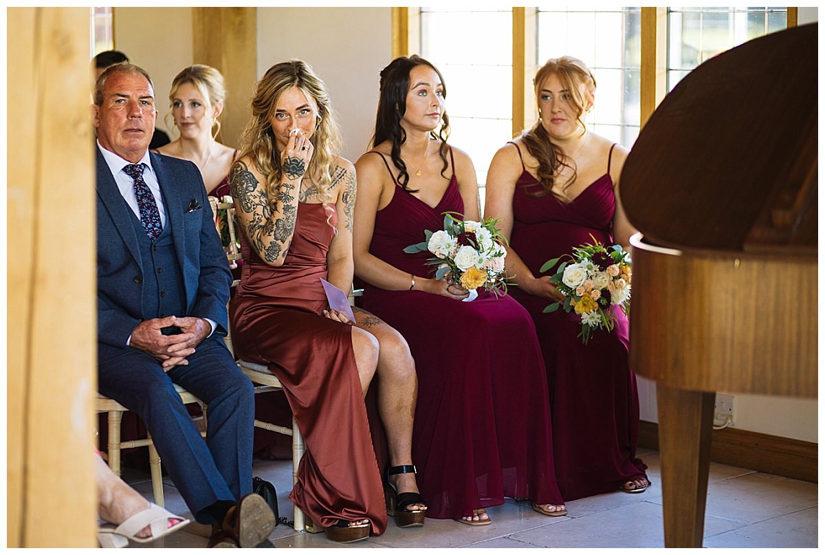 A group of guests, including bridesmaids and a guest in a suit, sitting in a venue, observed in a moment of anticipation during a wedding ceremony.