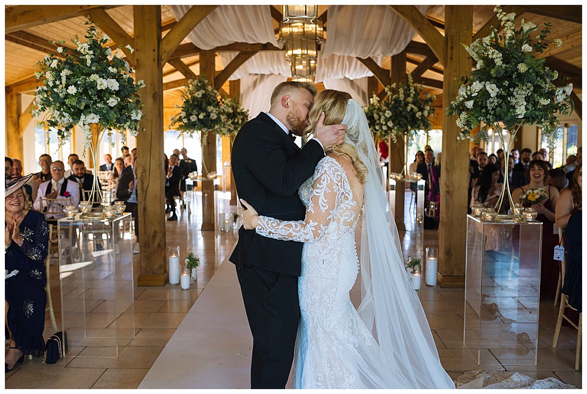 A bride and groom share a tender kiss during their wedding ceremony, surrounded by guests and elegant floral decor.