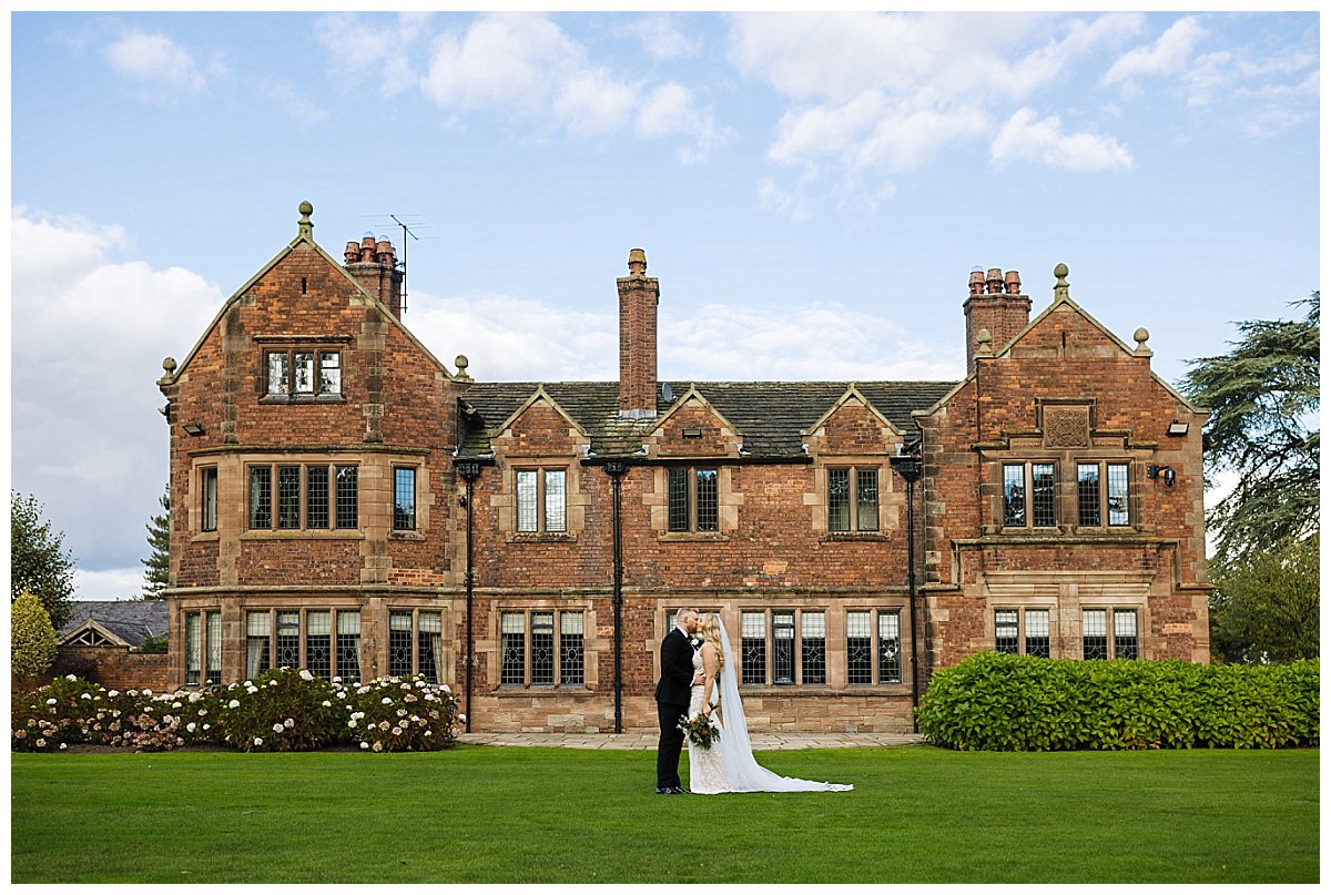 A bride and groom share a tender kiss on the lawn of a beautiful red-brick manor house, surrounded by lush greenery and blooming flowers under a partly cloudy sky.