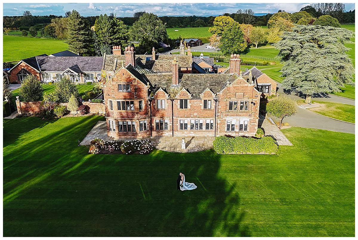 Aerial view of a beautiful brick manor house surrounded by greenery, featuring a couple posing on the lawn.