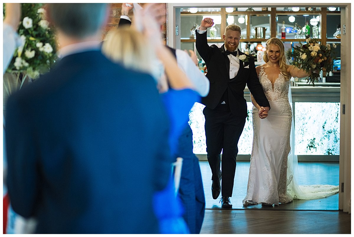 A smiling bride and groom joyfully enter their wedding reception, hand in hand, while guests celebrate with applause.