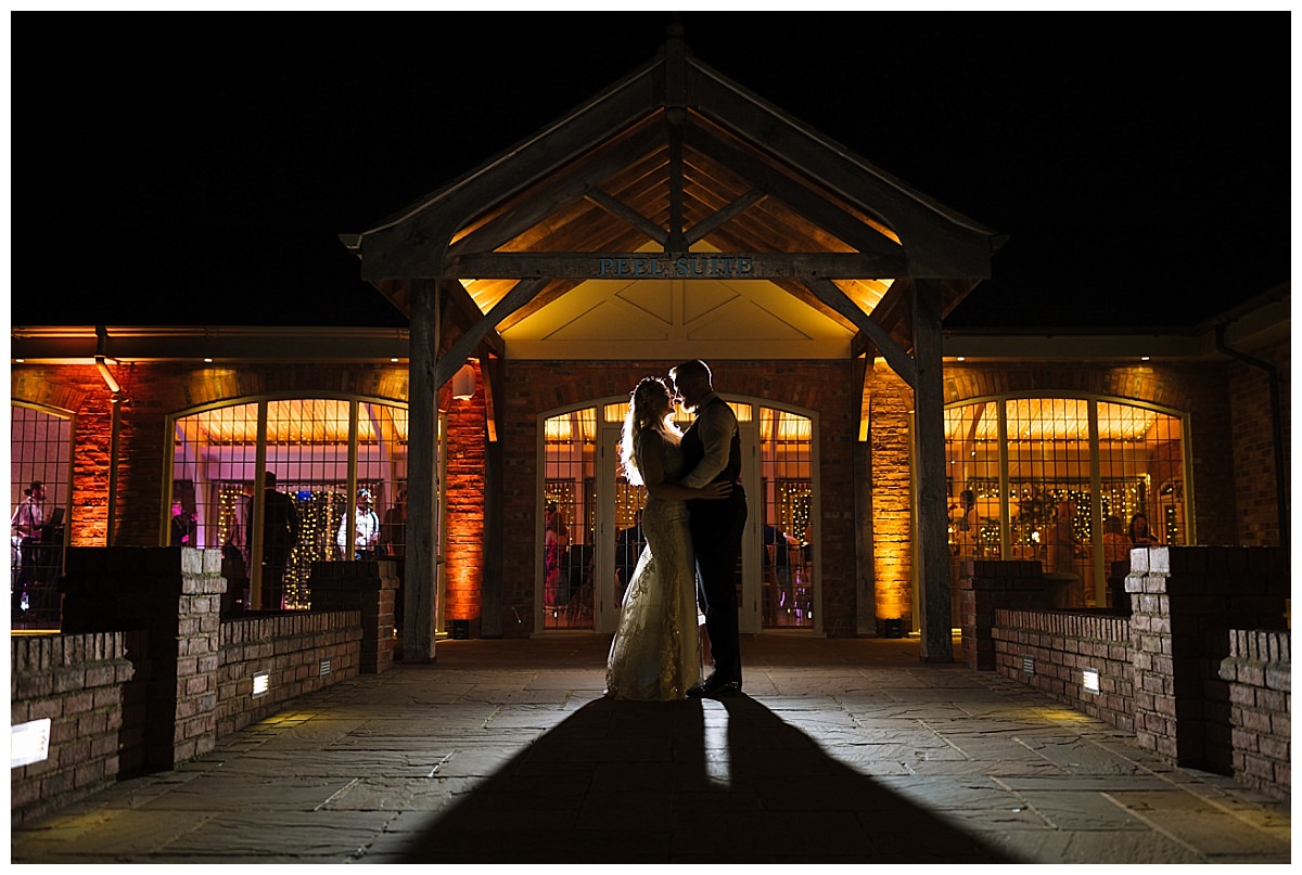 A couple shares a kiss in silhouette in front of a warmly lit venue at night.