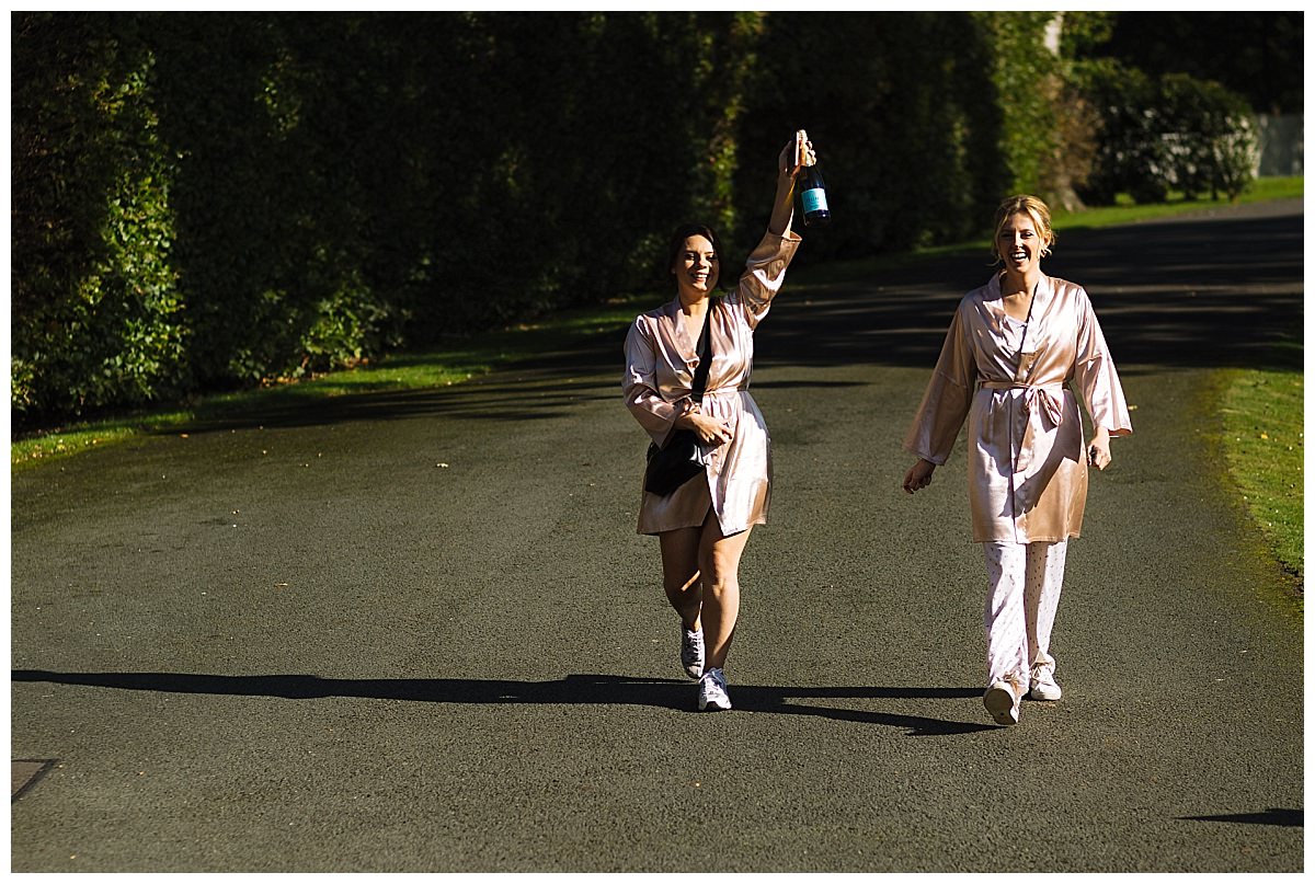 Two women walking down a quiet path at Colshaw Hall, dressed in matching silk robes, one holding a bottle in the air, exuding joy and celebration.