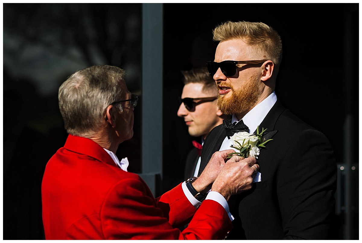 A man in a tuxedo receives a boutonniere from another man in a red jacket, while another guest in the background wears sunglasses at a black tie wedding at Colshaw Hall