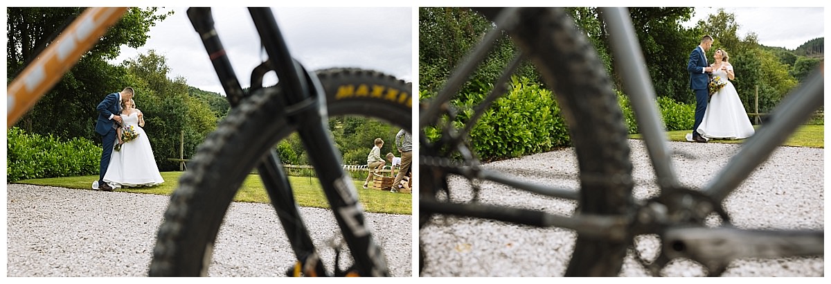 A couple in wedding attire embraces in a scenic outdoor setting, partially viewed through the frame of a bicycle.
