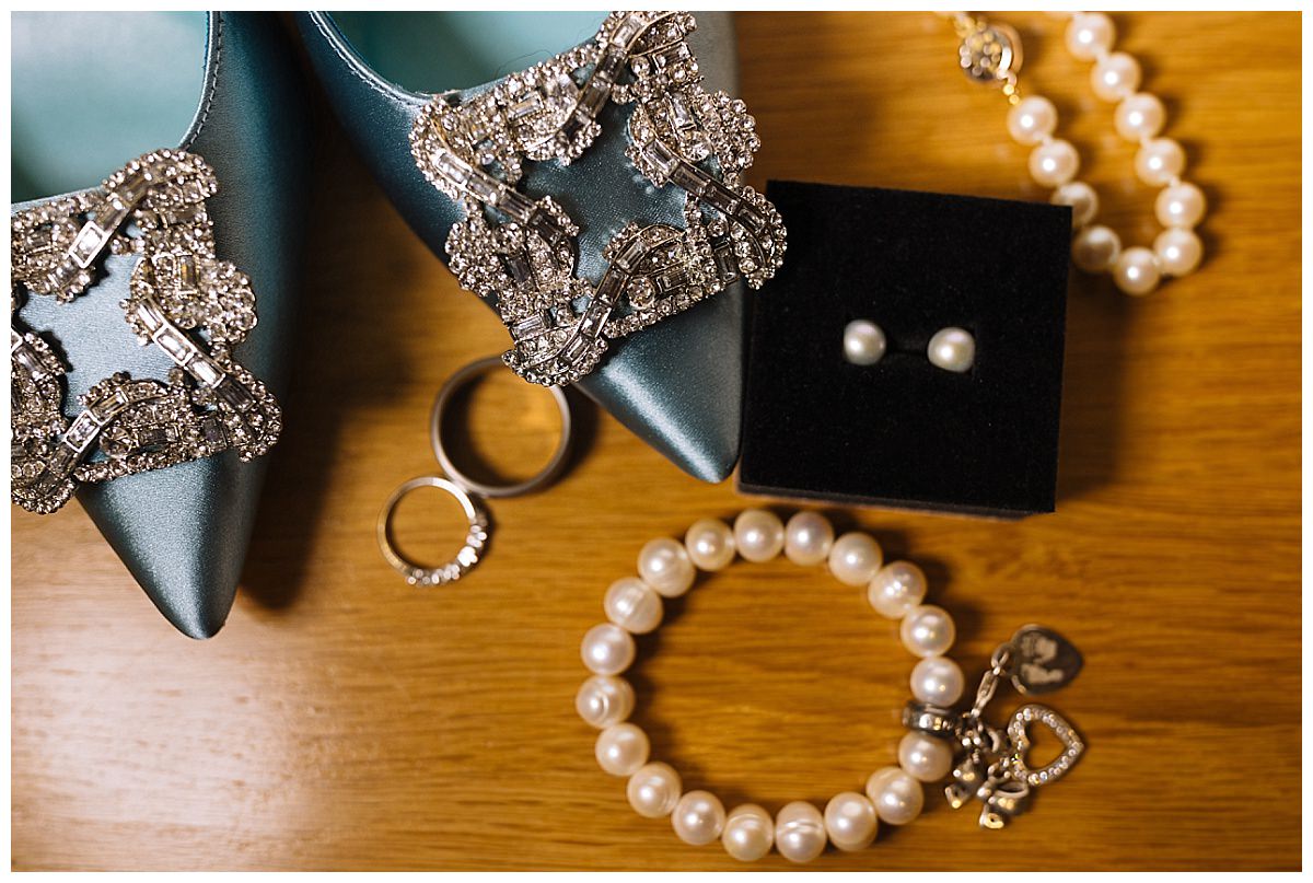 Close-up of blue satin heels with jeweled buckles, pearl jewelry, and engagement rings displayed on a wooden table.