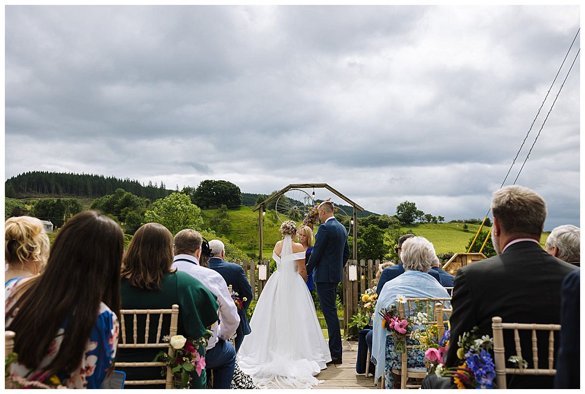 A bride and groom exchanging vows during an outdoor wedding ceremony, surrounded by guests in a picturesque landscape with greenery and cloudy skies.