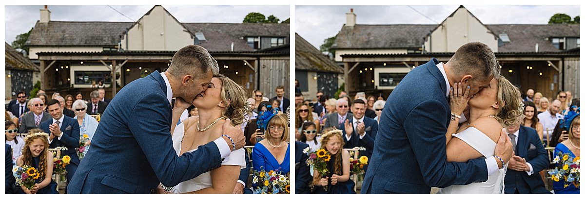 A newlywed couple shares a romantic kiss during their outdoor wedding ceremony, surrounded by a joyful audience of family and friends.