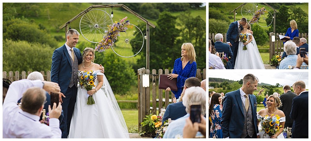 A joyful couple stands together during a wedding ceremony, surrounded by guests and scenic greenery, with a floral arch in the background.