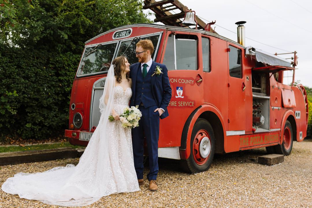 A bride and groom share a joyful moment in front of a classic red fire truck at Larkspur Lodge, showcasing a unique wedding theme.