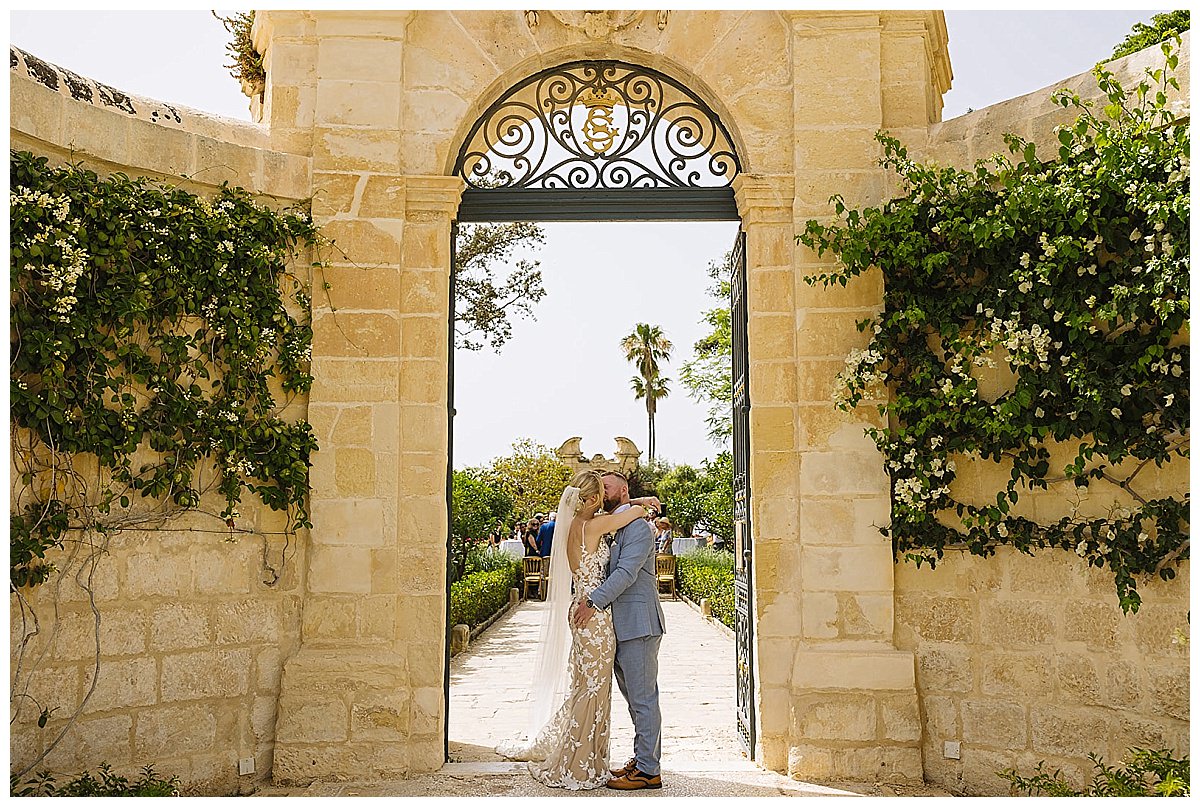 A couple shares a loving kiss under an ornate garden gate adorned with greenery, capturing a moment of joy on their special day.