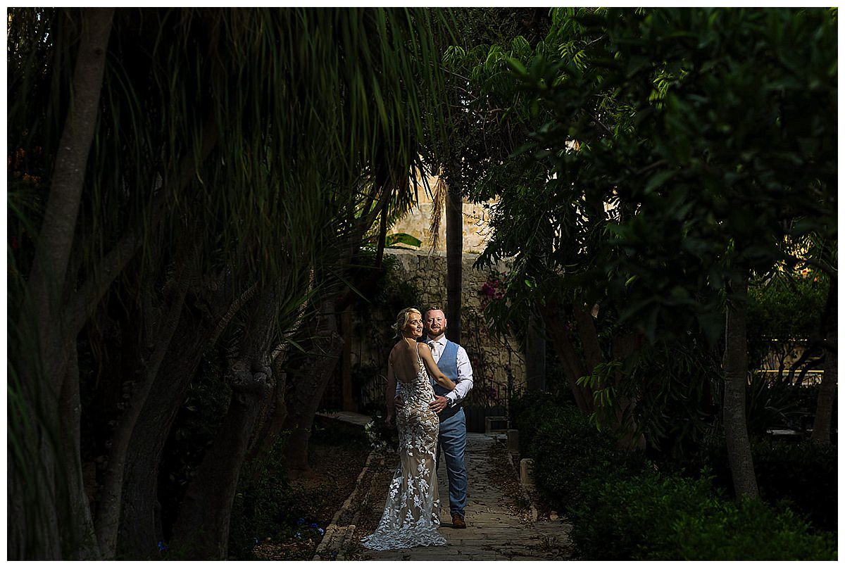 A couple in wedding attire shares a joyful moment in a lush garden at Palazzo Parisio in Malta, surrounded by trees and greenery.