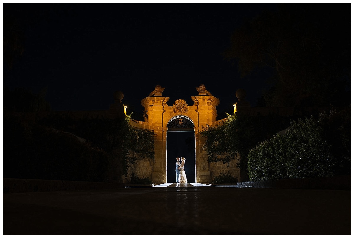 A couple shares a tender moment illuminated at a grand entrance of Palazzo Parisio, surrounded by lush greenery under a night sky.