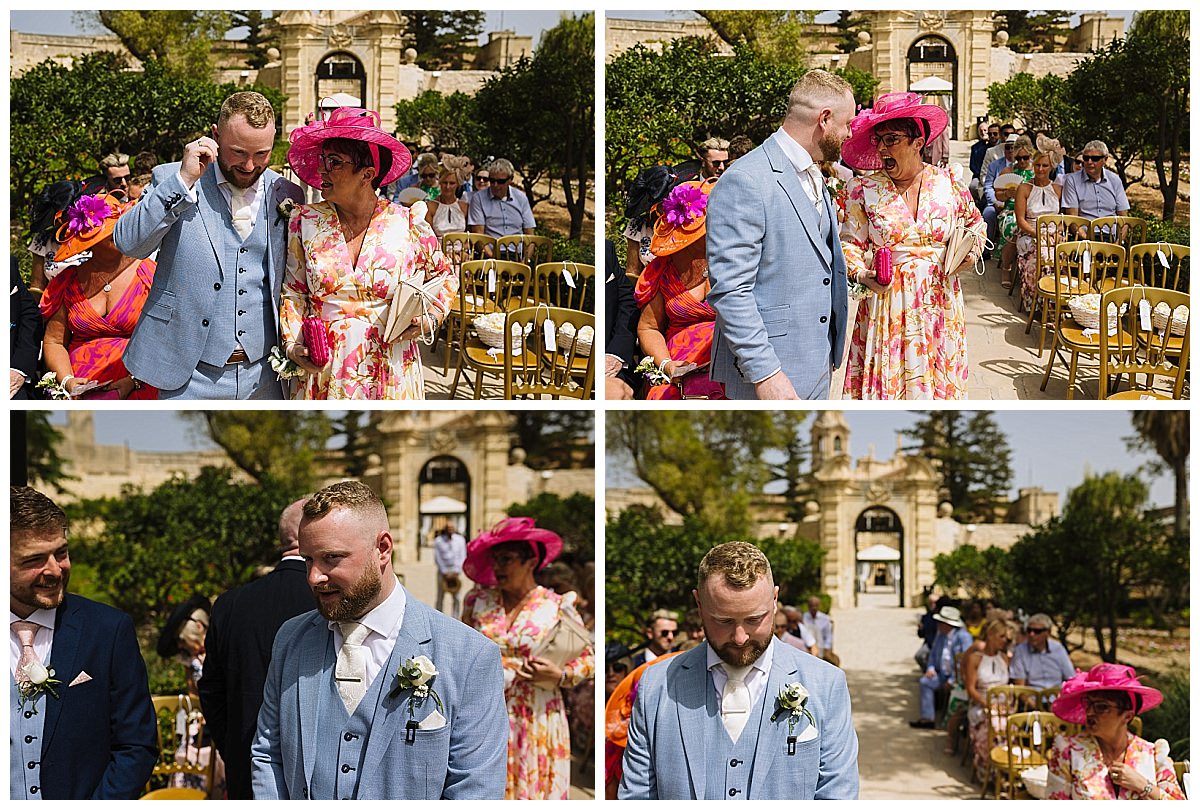 A groom in a light blue suit shares a moment with a woman in a floral dress and pink hat, surrounded by wedding guests in a scenic outdoor setting.