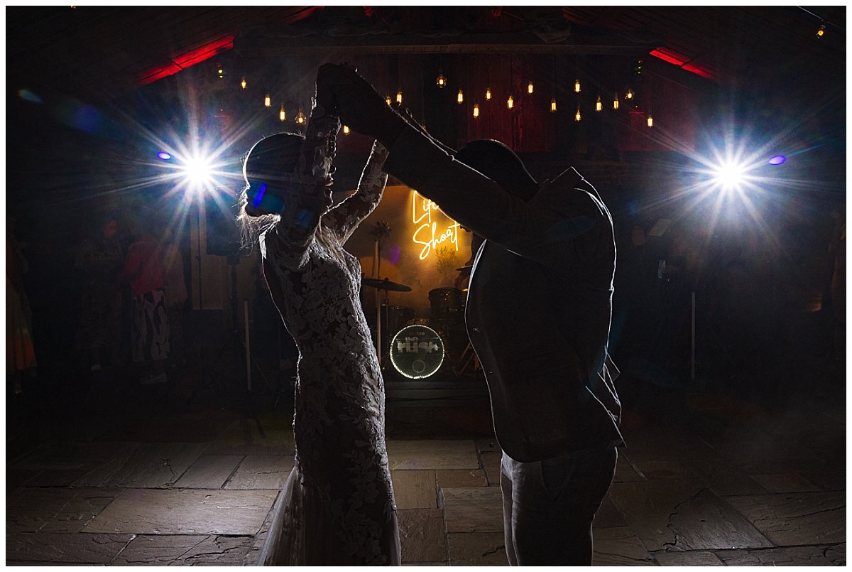 A couple joyfully dancing in a dimly lit venue with bright spotlights and a neon sign in the background, capturing a moment of celebration during wedding
