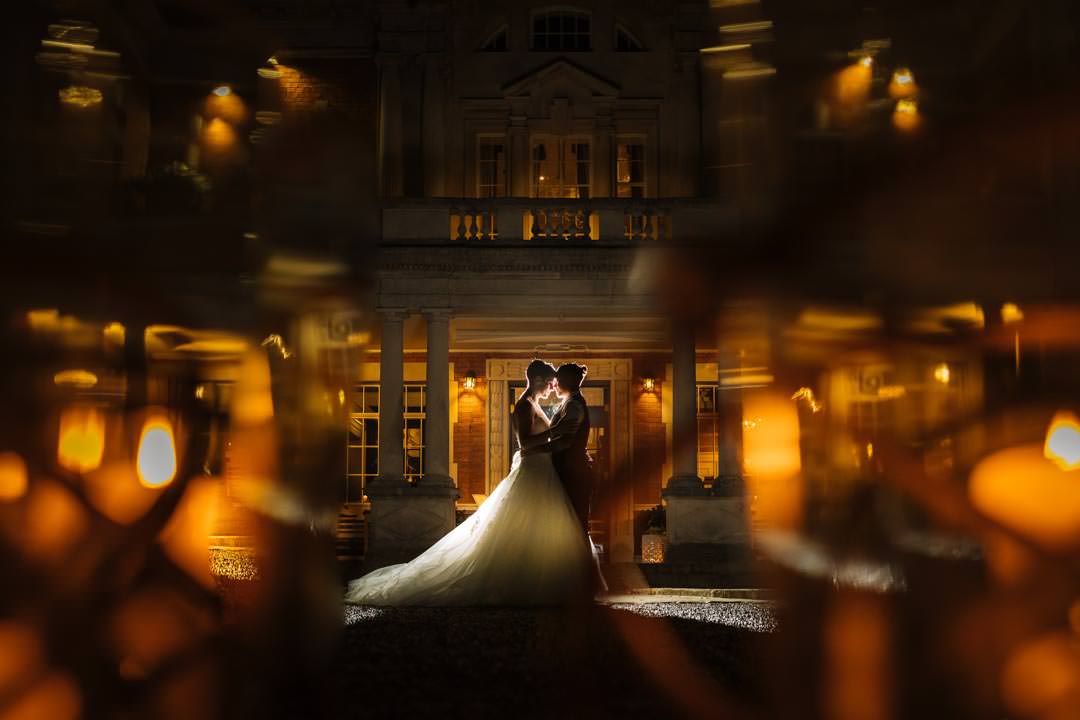 two brides embrace romantically in front of a beautifully lit eaves hall, captured in a dreamy atmosphere with warm, glowing highlights.