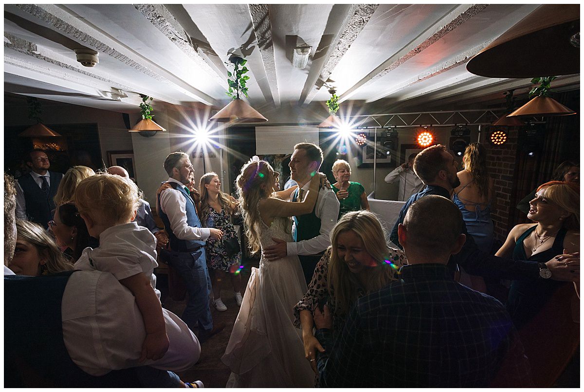 A joyful couple dances at their wedding reception surrounded by friends and family in a lively atmosphere with colorful lights.
