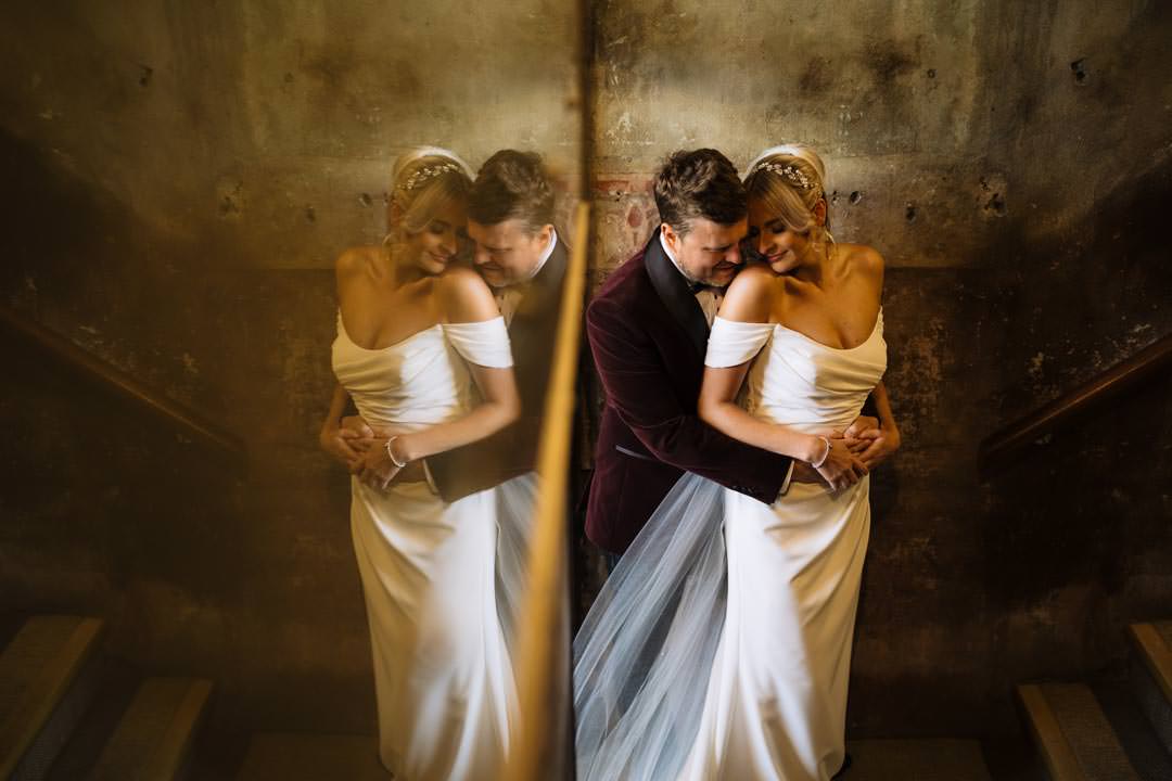 A bride and groom share a tender moment on a staircase, with reflections captured in the mirror at Holmes Mill showcasing love and joy on their special day.