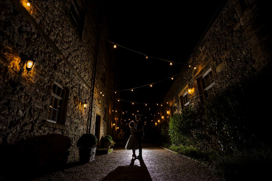 A couple dances in a beautifully lit courtyard surrounded by stone walls and twinkling lights at night.