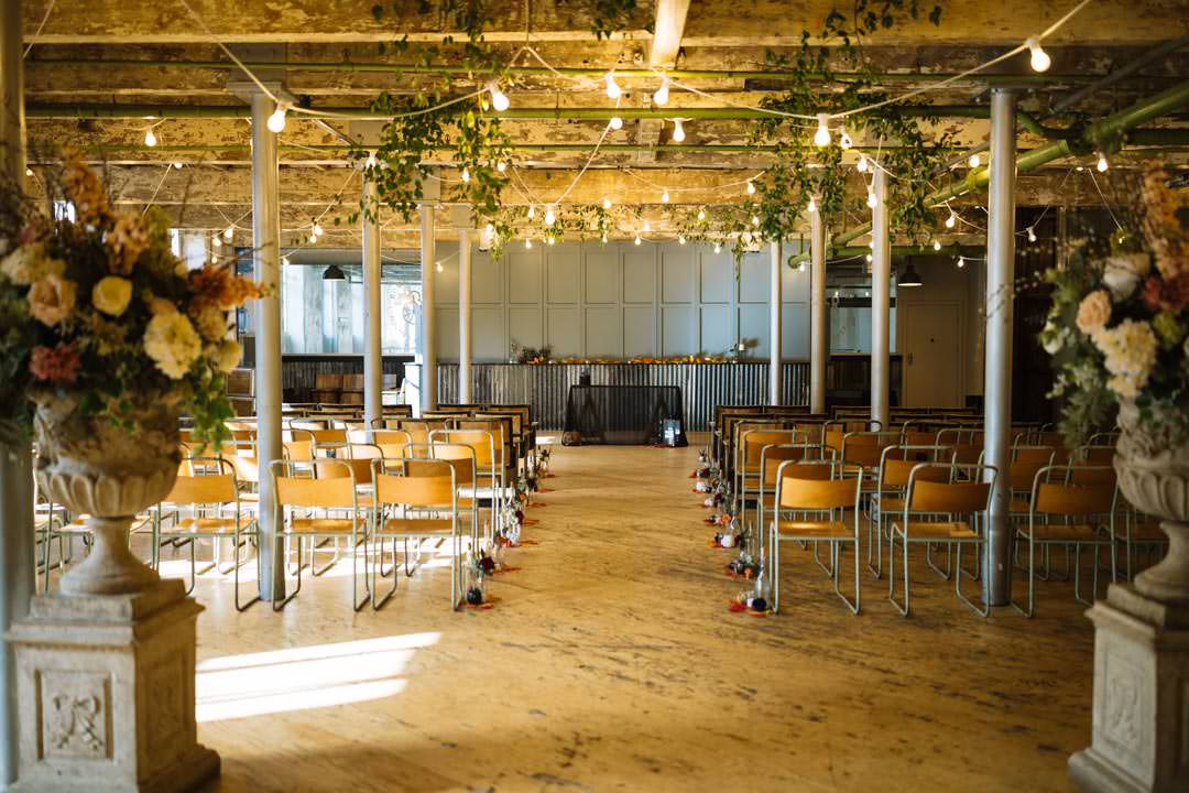 A beautifully arranged indoor venue featuring wooden floors, rows of chairs, decorative floral arrangements, and warm ambient lighting.