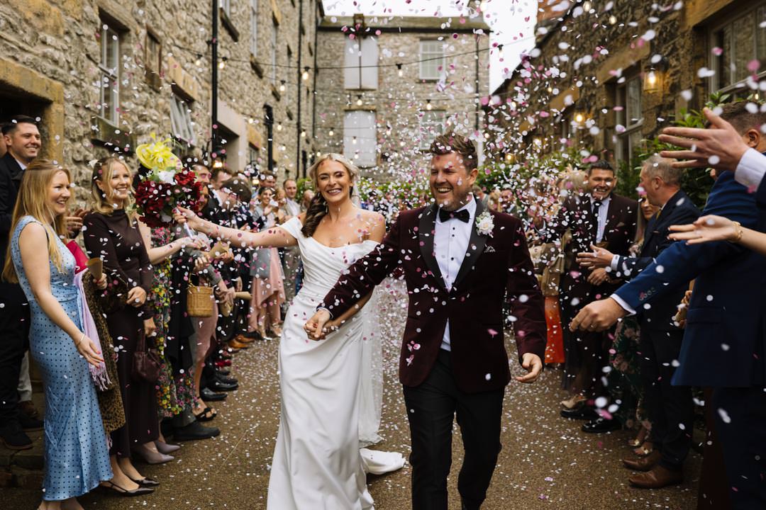 A joyful couple walks through a crowd celebrating their marriage, surrounded by colorful confetti at Holmes Mill