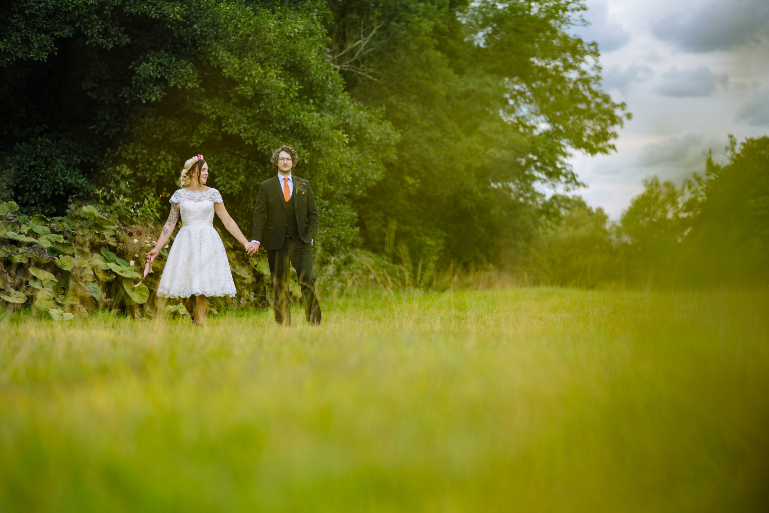 Couple Walking Hand-in-Hand in a Beautiful Landscape A happy couple dressed in wedding attire strolls hand-in-hand through a serene, green field surrounded by trees and lush foliage.