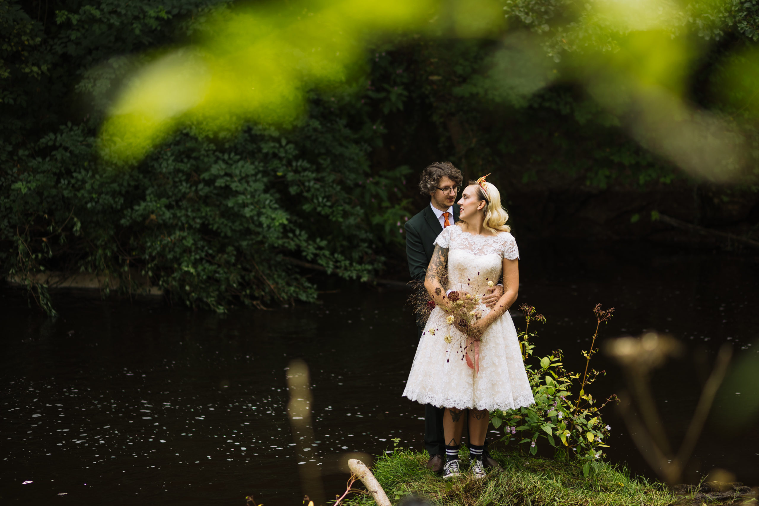 Romantic Forest Wedding Embrace A couple stands together by a serene river, the bride in a lace dress holding dried flowers, while the groom stands closely behind in a suit, surrounded by lush greenery.