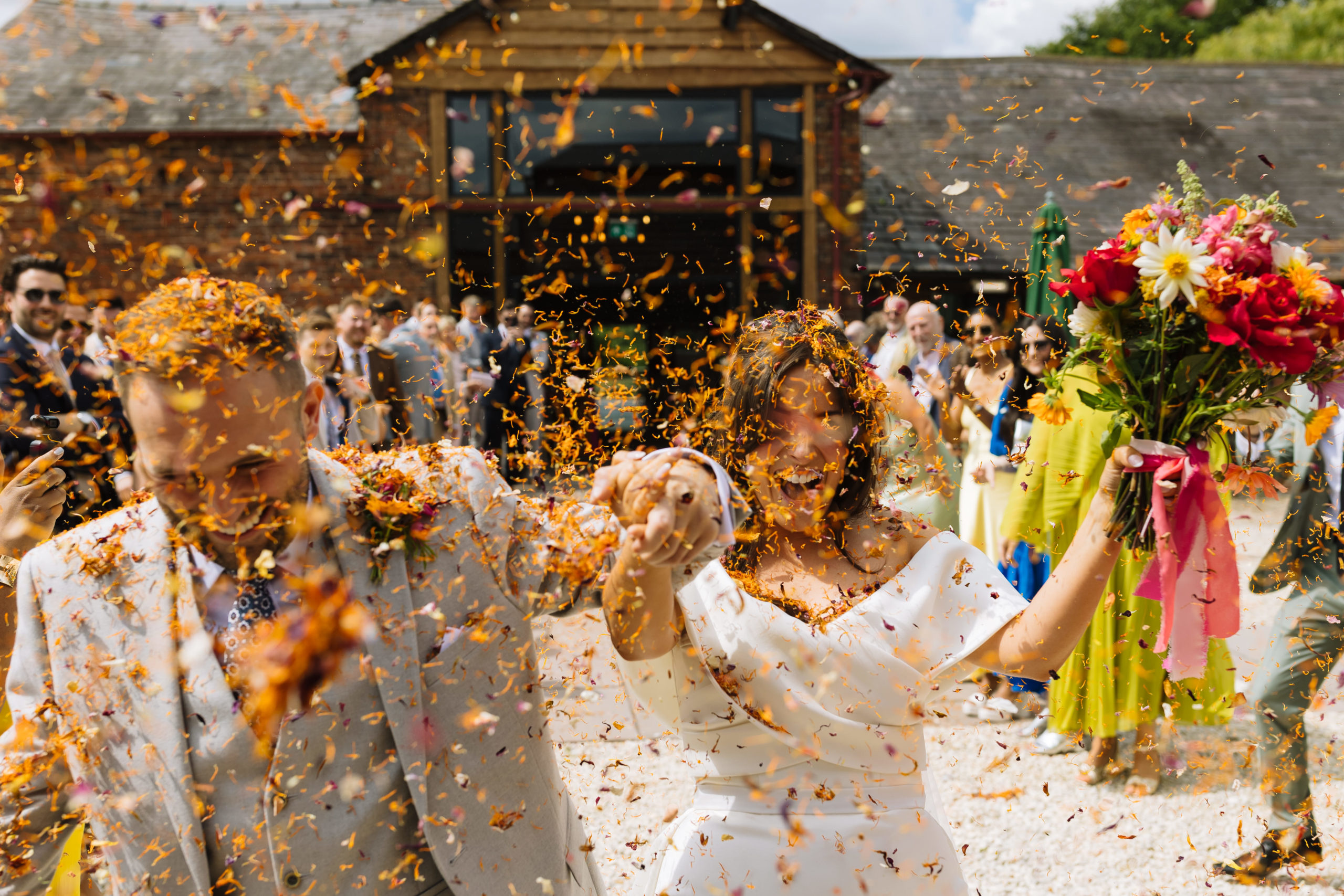 A joyful couple exits their wedding venue, covered in colorful confetti, while guests cheer in the background.