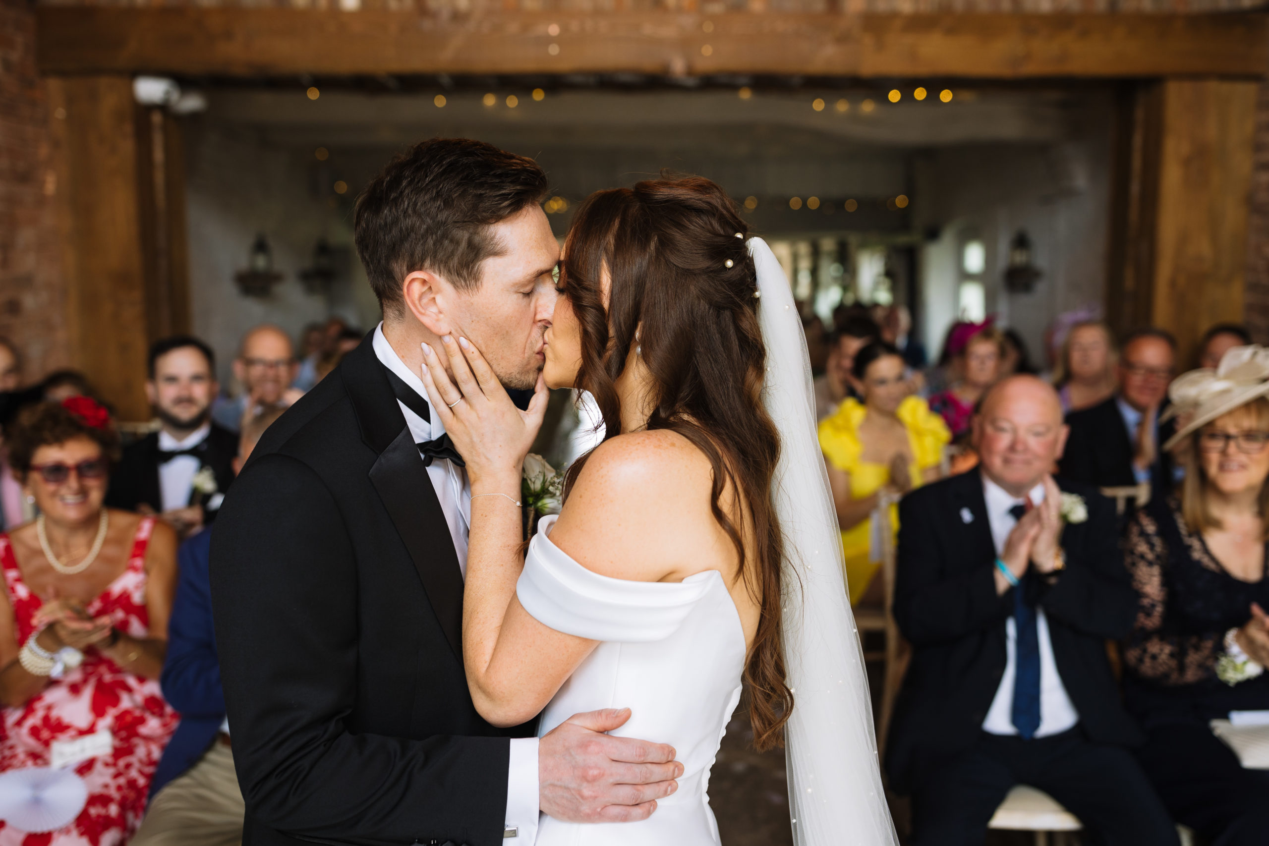 A bride and groom kiss during their Stock Farm wedding ceremony, surrounded by smiling guests applauding and celebrating the moment.