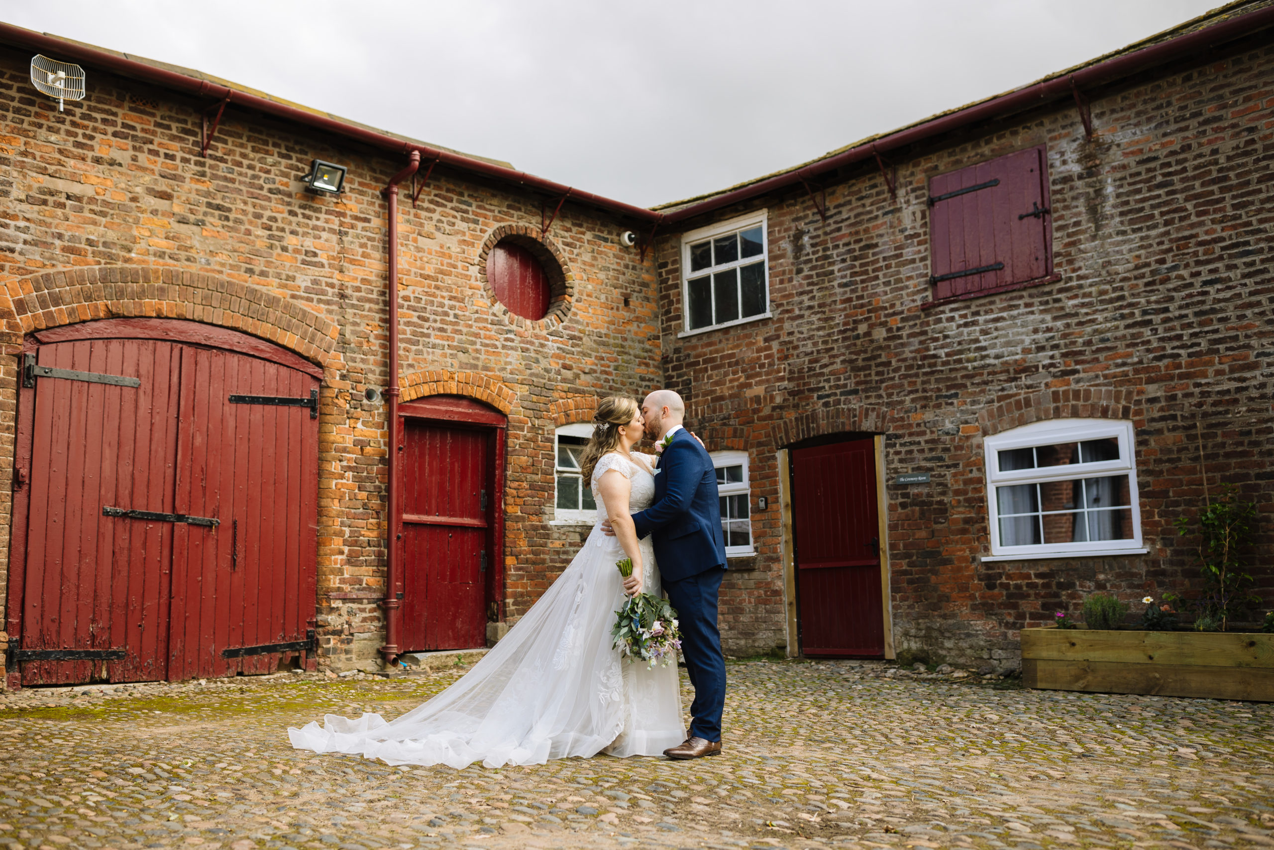 A couple shares a romantic kiss in front of a vintage barn with red doors, capturing a beautiful wedding moment.