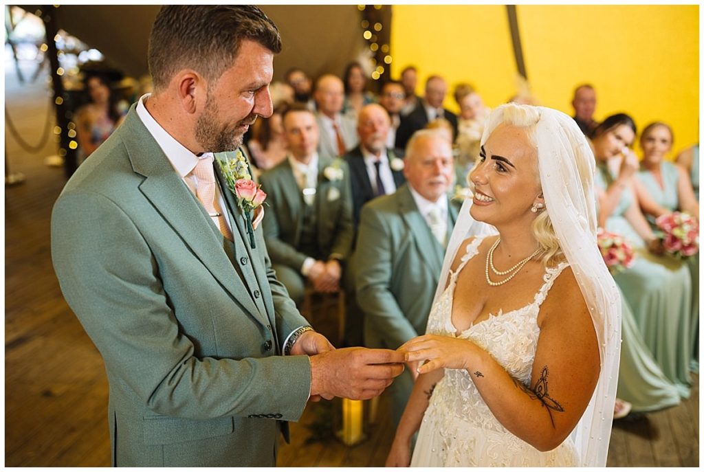 A bride and groom exchange rings during their wedding ceremony, surrounded by guests in an elegant setting.