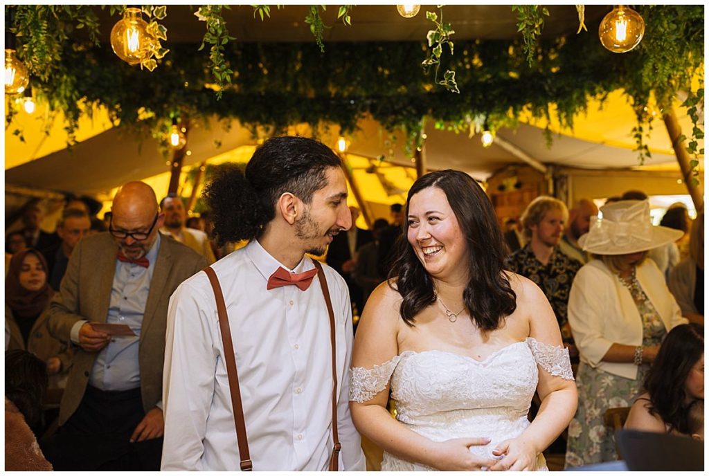 A bride and groom share a heartfelt smile while surrounded by guests in a festive, decorated setting with warm lighting and greenery.