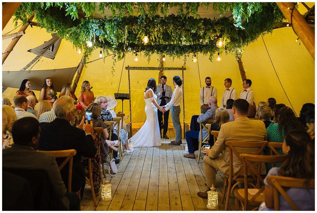 A couple exchanges vows during their wedding ceremony in a tent decorated with greenery, surrounded by family and friends.