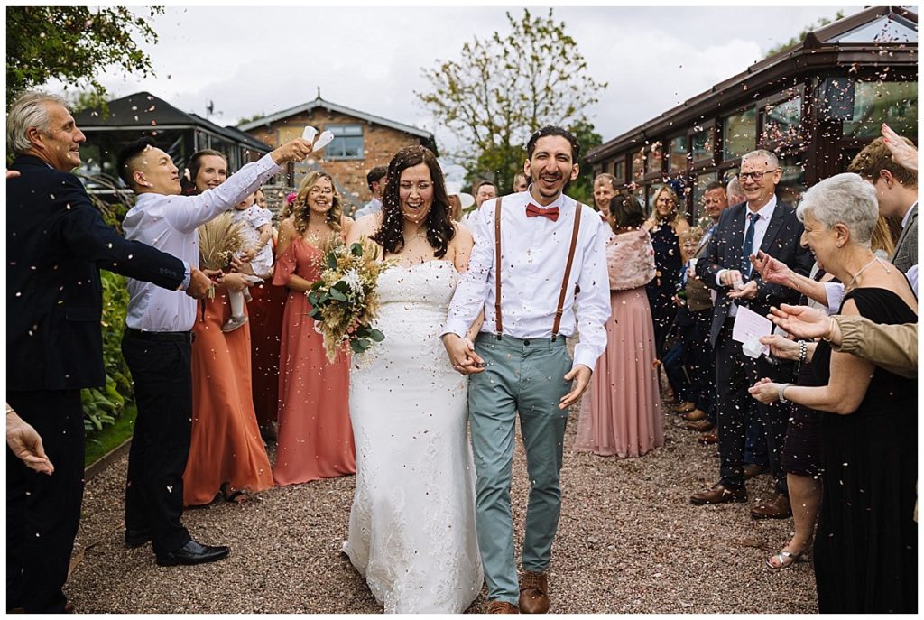 A newly married couple walks hand in hand, smiling and surrounded by guests throwing confetti in a picturesque outdoor setting.